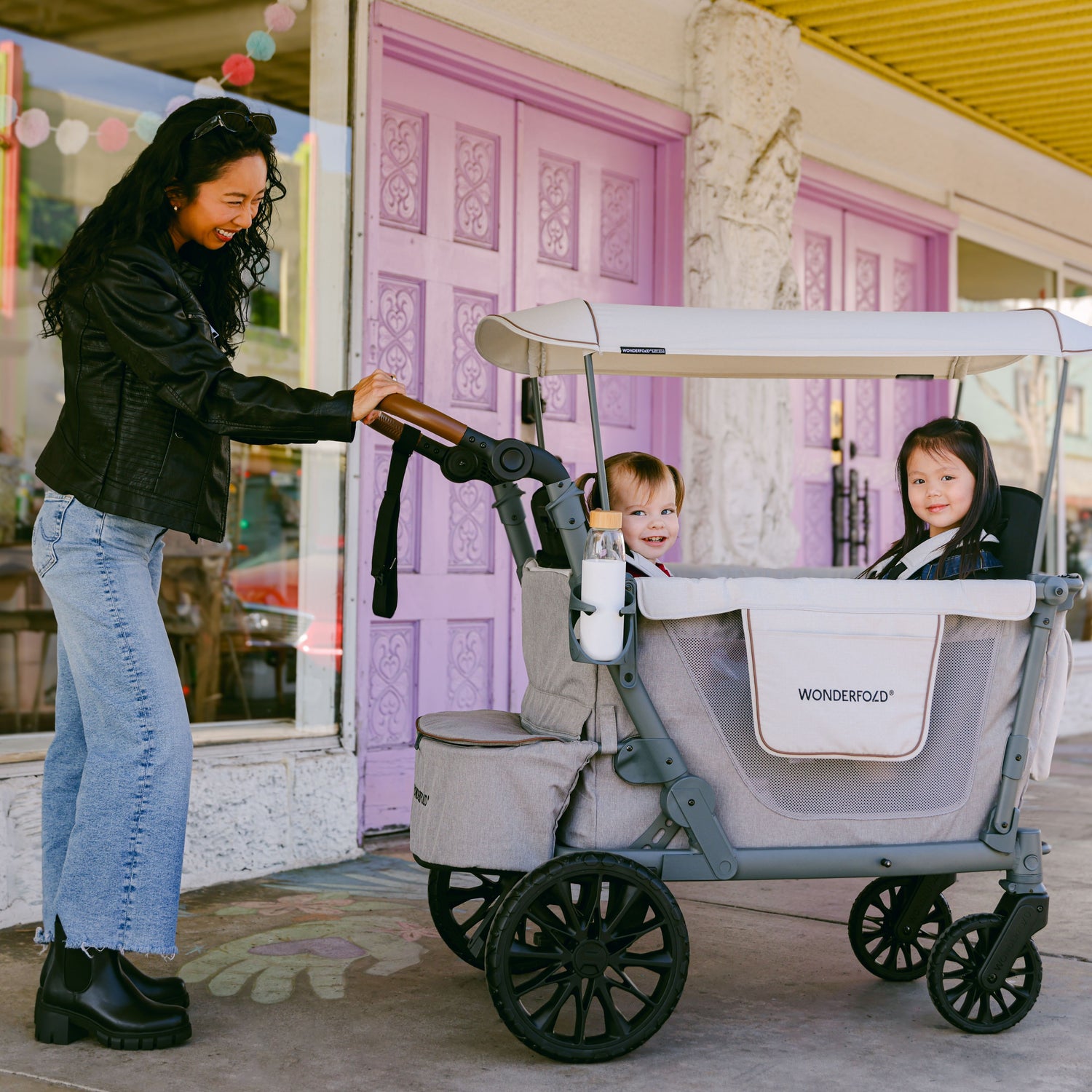 A woman pushing the WonderFold L2 stroller wagon with two happy children inside, strolling past a colorful storefront.